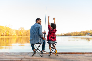An African American father and son sit on folding chairs on a wooden pier by a lake. The father, wearing a denim jacket, holds a fishing rod and looks at his son. The son, wearing a red plaid shirt