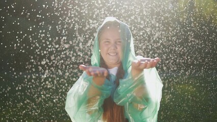 A child in a raincoat stands under raindrops. Happy family kid dream concept. Girl in a raincoat is raining and smiling. The girl heavily in the raincoat is smiling, it is raining.