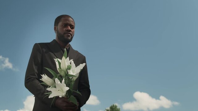 Sad African American man dressed in black suit holding bouquet of lilies outdoors under clear blue sky at funeral service