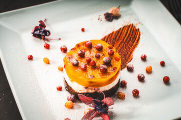 Dessert or cake made of mango, sea buckthorn and lingonberry jelly with cottage cheese or cream and chocolate on a white plate decorated with flowers, berries and chocolate.
