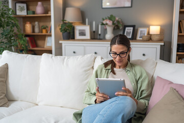 Relaxed young adult caucasian woman read a book on modern device reader comfortably sitting on the couch at home Portrait of pretty female people wearing eyeglasses and using digital tablet connection