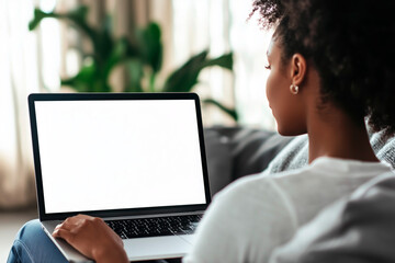 An over-the-shoulder view of an African American woman working on a laptop with a blank screen, seated comfortably in a bright, modern living room. Ideal for remote work, technology, and lifestyle con