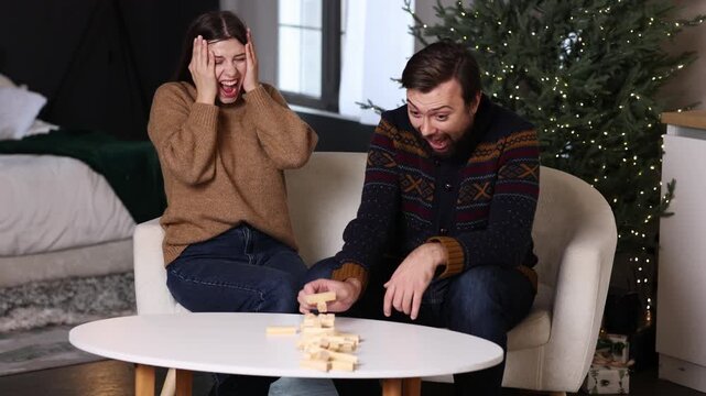 woman looks terrified while man mischievously after pulling block from a Jenga tower. woman's hands are over her face. man holding block hand. fun and board games on winter weekends Christmas tree 