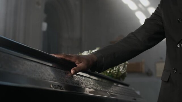 African American man in black suit passing his hand over coffin lid with white flowers at funeral service in cathedral
