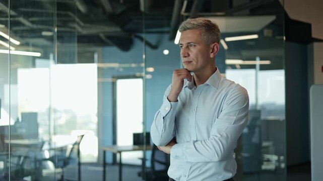 Thoughtful male business person thinking about work standing by window in a modern office. Pensive professional thinking in office. Focused business man face looking forward. Serious man portrait.