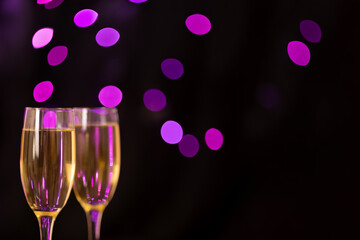 Champagne glasses overflowing with bubbles, positioned on the left side with a black backdrop and out-of-focus violet lights, ideal for a festive occasion.