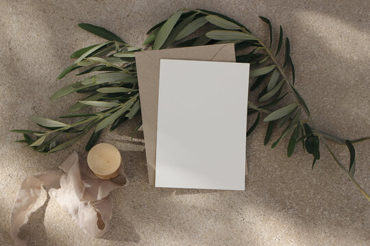 Italian summer wedding stationery mock-up. Blank vertical greeting card, invitation with envelope. Olive branches, silk ribbon on beige limestone background in sunlight. Moody flat lay, top view.