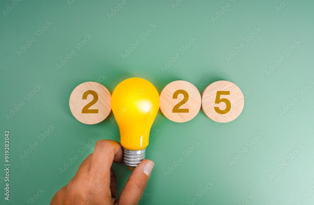 Wall mural Happy new year 2025 innovation technology, creative market trend, inspiration and idea concept. 2025 calendar number on wood blocks with glow yellow light bulb holding in man hand on green background.