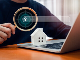 Online searching property, house value, home buy and sell, real estate investment concept. Location map pointer icon in magnifying glass in man hand with search engine bar near white house on laptop.