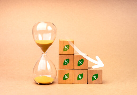 Arrow down on wood cube blocks as graph steps with green leaf with percentage icon near hourglass, isolated on brown background. Eco-friendly business with sustainable environment care concept.