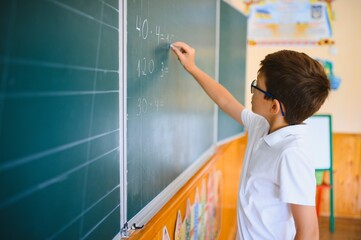Cute little schoolboy diligent write chalk solve math problem dressed stylish uniform classroom blackboard background