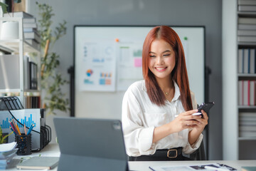 Asian businesswoman is sitting at her desk and smiling while using her smartphone