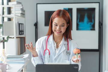 Smiling doctor explaining using anatomical brain model while having video call on tablet in clinic office