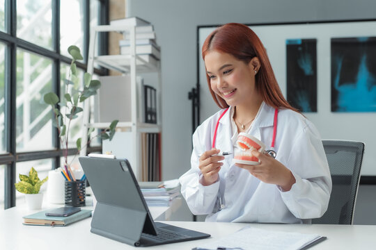 Asian female dentist is explaining teeth anatomy to her patient using a digital tablet and a denture mold during an online consultation