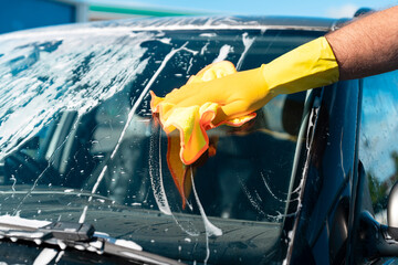 Person in yellow gloves wipes down  car windshield, removing soap bubbles under bright blue sky