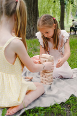 Preschool girls carefully stacking wooden blocks while playing tower game on blanket in a park filled. Attention and informal education