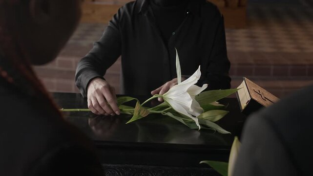 Unrecognisable woman putting white lilies on coffin lid during funeral service held by priest in church