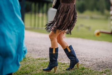 Riga, Latvia - August 10, 2024 - Close-up of a person wearing blue denim boots and a brown fringed...