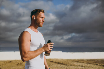 Young cool strong athletic toned fit sportsman man wears sports clothes earphones drink protein...