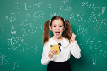 Little school girl 7-8 years old pony tails wear white shirt uniform hold use mobile cell phone do winner gesture isolated on green blackboard background. Childhood kids education lifestyle concept.