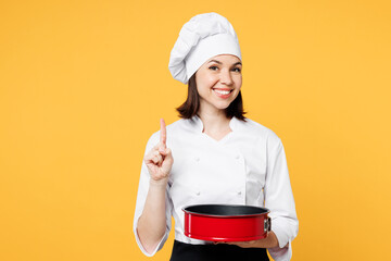 Young chef cook baker woman in white shirt black apron uniform toque chefs hat holding red non-stick baking form for pie cake point finger up isolated on plain yellow background Cooking food concept