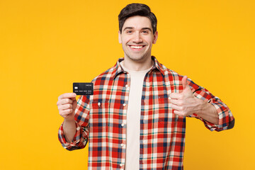 Young smiling happy man wears red checkered shirt casual clothes hold in hand mock up of credit bank card show thumb up isolated on plain yellow orange background studio portrait. Lifestyle concept.