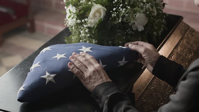 Close up shot of unrecognisable female relative putting American flag folded in triangle on coffin lid next to white flowers during memorial ceremony in church