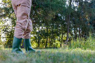 Woman gardener in brown garden overalls and rubber boots on the garden in warm autumn sunny day. Lush vegetation and grass and trees background. Autumn gardening.