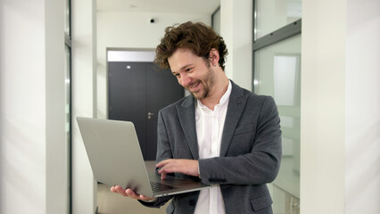 Confident young professional in a modern office corridor, using a laptop and displaying self-assuredness in his career and work.