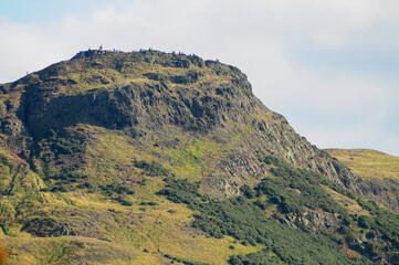Hikers atop a rugged, grassy peak in Edinburgh, Scotland, enjoying scenic views.