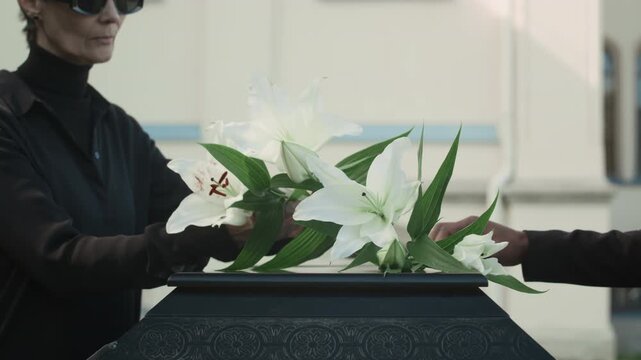 Unrecognisable Black woman and Caucasian lady putting white lilies on coffin lid during outdoor funeral service