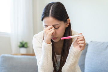 Sad, Disappointed motherhood asian young woman holding pregnancy test, upset ovulation problem, anxiety after result at positive, negative sitting on couch at home. Unwanted, contraception concept.