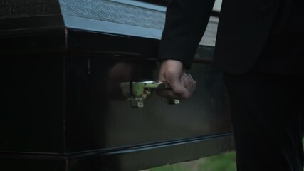 Close up view of hand of unrecognisable young man dressed in black suit holding coffin handle and carrying it to cemetery