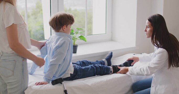 Cute little child boy sitting on couch with mother in medical clinic with broken leg in plaster during check up in exam room. Friendly doctor traumatologist giving consultation in hospital. 4k video.