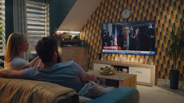 Adult couple talks and watches news broadcast report about US Election Day on the big TV screen sitting on sofa at home. TV display shows playback of breaking news with female Caucasian reporter.