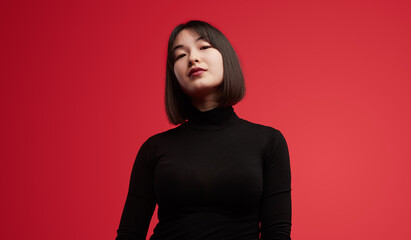 A confident woman wearing a black turtleneck poses against a vibrant red background, exuding...