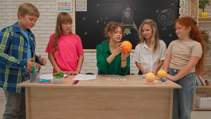 Junior school boys and girls sit in room in astronomy class. Female teacher holds planet model and...