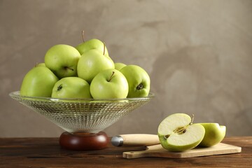 Glass vase with fresh green apples, cutting board and knife on wooden table against gray background