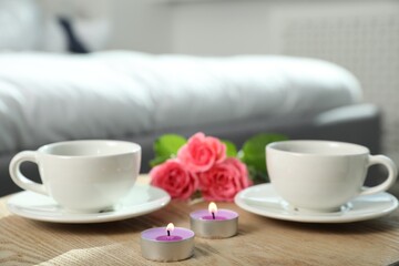 Burning candles, rose flowers and cups of drink on wooden table indoors