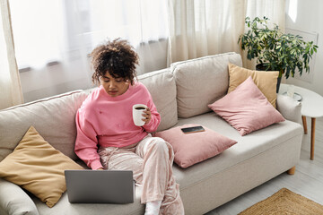 A woman relaxes indoors, working on her laptop with a warm drink in hand.