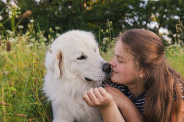 Beautiful young woman kissing her cute large white Great Pyrenees dog laying in tall grass meadow, enjoying time together. Pet owner, pet love, dog frienship concept.