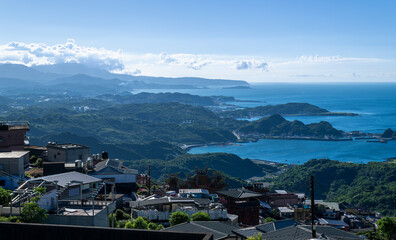 Fototapeta premium A scenic view of Jiufen Village with green mountains stretching to the blue sea, under a blue sky with white clouds. The picturesque afternoon landscape. Jiufen, Taiwan.