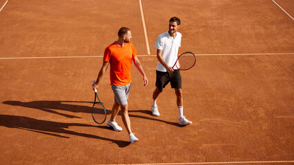 Two men, tennis players walking on outdoor clay court with racket, discussing strategy of the game. Team training and competing. Concept of sport, competition, active and healthy lifestyle