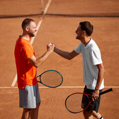 Two smiling men, tennis players on clay court exchanging handshake before game session, showing teamwork and readiness for good training. Concept of sport, competition, active and healthy lifestyle