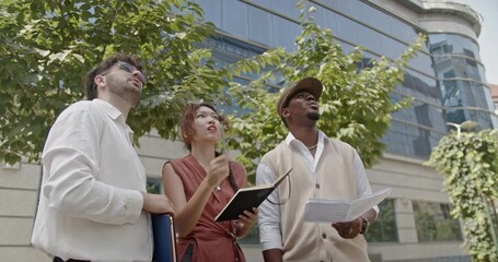 Three professionals engage in an outdoor discussion while observing a building under construction. They hold documents and notebooks, sharing insights and ideas, emphasizing their collaborative