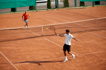 Two men, focused tennis players playing fast-paced tennis match on outdoor clay court, showing ambitions and skills during training session. Concept of sport, competition, active and healthy lifestyle