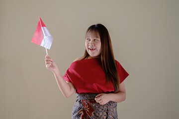 An excited Asian woman wearing a red dress and traditional sarong is celebrating Indonesian Independence Day by holding an Indonesian flag on an isolated background