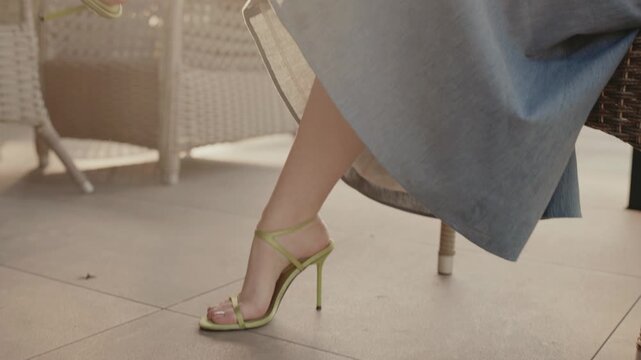 Close-up of a woman's legs wearing green high heels and blue dress while seated on an outdoor patio. Close-Up of Woman's Legs in Stylish Heels and Dress on Outdoor Patio