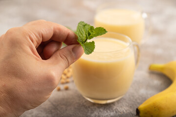 Organic non dairy banana and soy milk in glass with hand on brown concrete, side view, close up, selective focus.
