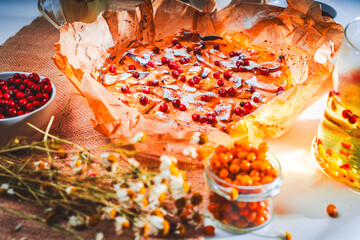 Apple tartate decorated with lingonberries with herbal and berry tea on a white table with sea buckthorn in a bowl and dried chamomile on a canvas napkin